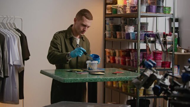 Medium stab shot of Caucasian male designer in gloves handling blue paint with spatula during preparation for branding and screen printing job on marble worktable in workshop