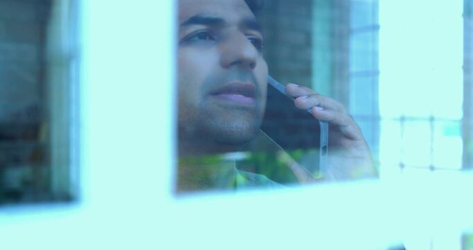 Indian impatient young man distressed because routine call fail using smartphone standing near glass window in modern Indian home with view from outside showing reflections on the glass pane