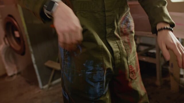 Tilt down shot of Caucasian male worker putting his smartphone in pocket of his coveralls and starting browsing stack of screen printing frames in creative workshop, preparing for branding job