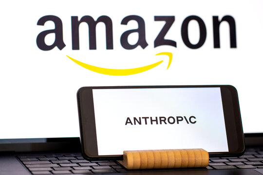 Anthropic logo is displayed on a mobile phone in the foreground, while the Amazon logo appears in the background on a computer screen. Anthropic is an artificial intelligence company, and Amazon is