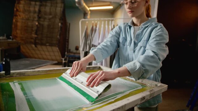 Eye level medium shot of young woman applying ink on screen frame using squeegee in workshop environment, showing precision, branding process and creative industry work