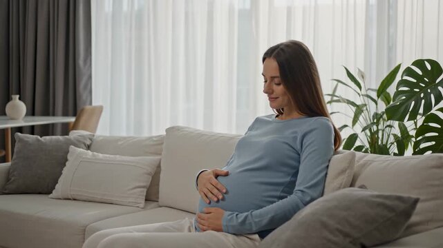 Pregnant woman sitting on sofa at home gently holding her baby bump, maternity wellness and expecting mother relaxation in modern living room