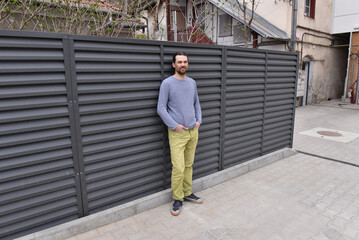 Fototapeta premium Man in grey sweater and yellow pants leaning against a dark louvered metal privacy fence