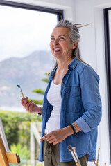 Naklejka premium Senior woman standing in bright art studio holding paintbrush with green paint, denim shirt