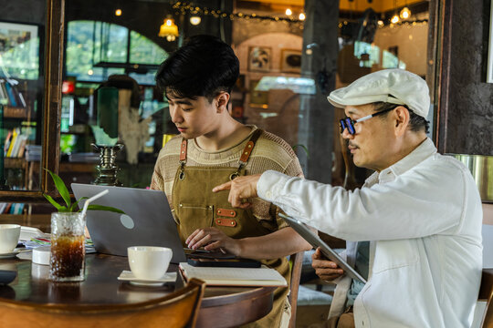 Modern small business digital management young entrepreneur using laptop and tablet planning, finance workflow efficiency technology cafe business operation, learning from senior cafe owner experience