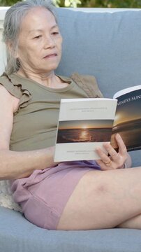 Vertical video: Smiling senior woman reading sunset-cover book on porch, tracing lines in olive top