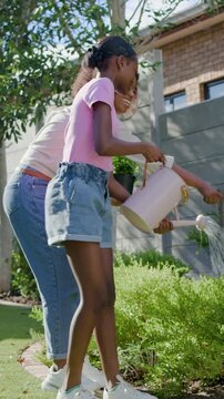 Vertical video: Approaching mom w or  pot guiding girl in pink T, tipping can, watering shrubs in ya