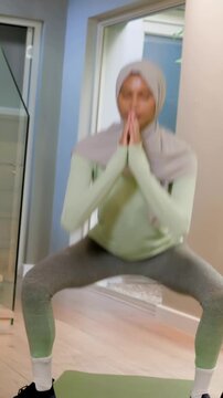 Vertical video: Starting workout, woman in hijab doing sumo squats in hall with kettlebell