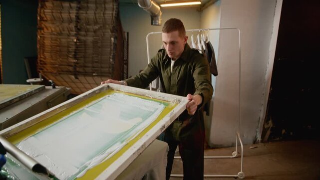 Medium shot of young male worker operating squeegee on screen printing frame installed on press in workshop, showing manual technique and creative production
