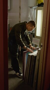 Vertical shot of Caucasian male worker slipping his smartphone into his coveralls pocket and beginning to sort through stack of screen printing frames in creative workshop