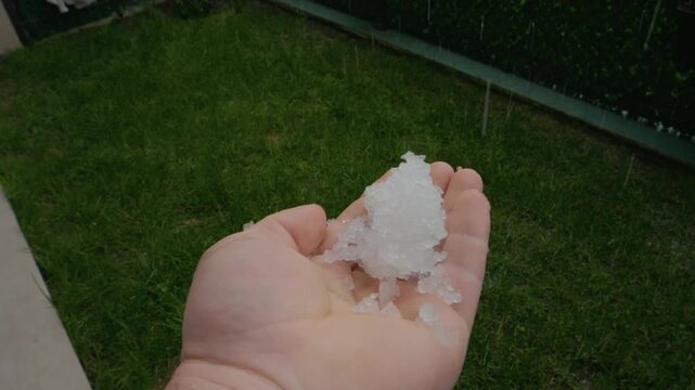 A man shows chunks of ice in his hand as hail falls from the sky. Heavy rain accompanies the storm, creating a wet and icy scene outside.