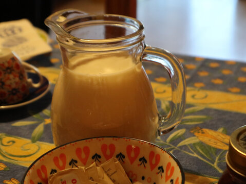Caraffa in vetro trasparente piena di latte fresco sul tavolo da colazione, primo piano con ciotola decorata e tovaglia in stile rustico mediterraneo per la ristorazione e l'ospitalit&agrave;
