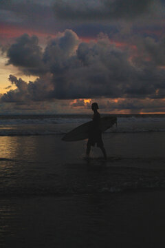 Lone surfer silhouette leaving ocean under moody evening sky, Bali