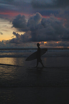 Surfer with board walking along shoreline at colorful dusk, Bali