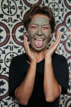 Playful woman with green clay face mask sticking tongue out, Bali