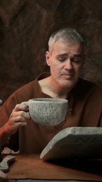 Funny sleepy prehistoric man having breakfast in his cave: he is reading news and drinking a big cup of coffee