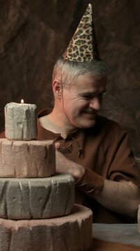 Cheerful happy prehistoric man celebrating his birthday in his cave, he blows the candle on a huge birthday cake
