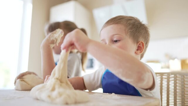 Child kneading dough with focused hands, boy and girl toddler nearby, play and baking activity in bright kitchen at home, family interaction around dough, hands covered in flour and smile and joy