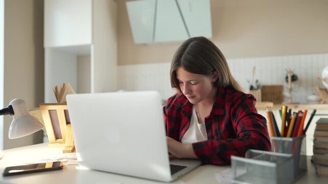 Teen studies on laptop at desk, young girl focused on homework and study tasks, student leaning toward screen, learning at home with book stack and pencil holder showing concentration and education