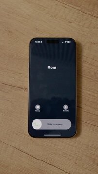 Vertical Screen: Smartphone sits on a wooden table. An incoming call appears with Mom's name on the screen. The time is shown at the top of the phone. A simple moment at home.