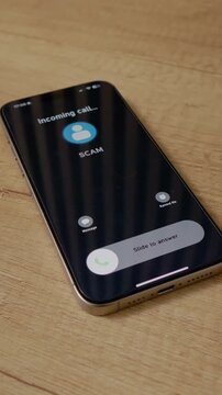 Vertical Screen: A smartphone receives an incoming call labeled as Scam while placed on a wooden table. This moment raises awareness about phone scams.