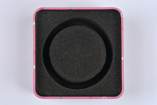 Top view of an empty square pink metal watch box with a circular black foam protective insert on a light background
