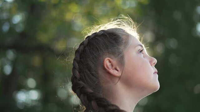girl breathing in warm sunlight, eyes closed, profile turned upward among green tree bokeh, braid and hair illuminated, peaceful child portrait conveying relaxation and nature connection outdoor