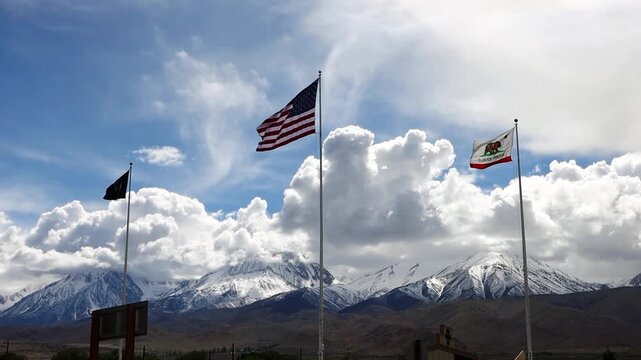 American, California, and MIA POW flags during a wind advisory warning in the eastern sierra and Owens Valley, With blue sky and puffy clouds in background.