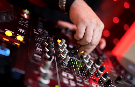 DJ Mixing Music on Professional Controller in Nightclub. Close-up of a DJ's hand adjusting knobs and faders on a professional mixing console. Blurred red bokeh lights in the background