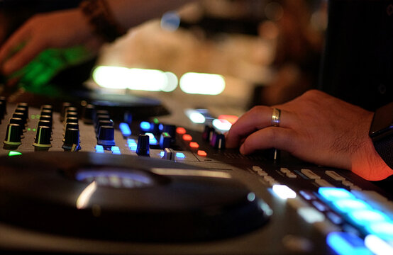 DJ Mixing Music on Professional Controller in Nightclub. Close-up of a DJ's hand adjusting knobs and faders on a professional mixing console. Blurred red bokeh lights in the background