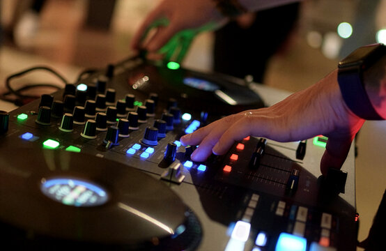 DJ Mixing Music on Professional Controller in Nightclub. Close-up of a DJ's hand adjusting knobs and faders on a professional mixing console. Blurred red bokeh lights in the background