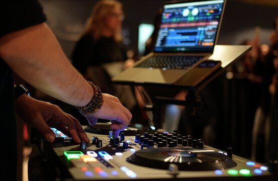 DJ Mixing Music on Professional Controller in Nightclub. Close-up of a DJ's hand adjusting knobs and faders on a professional mixing console. Blurred red bokeh lights in the background