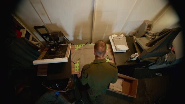 Top view of worker folding t shirts on table near printing equipment and putting them inside cardboard box, showing packaging stage and apparel workflow