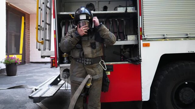 Professional firefighter putting oxygen mask against background of big truck. Young fireguard in full equipment near fire engine ready to work. Concept of saving lives and heroic profession. Slow mo