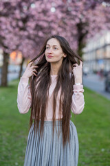 Naklejka premium Woman with long brown hair wearing sunglasses poses among blooming sakura cherry blossom trees in a vibrant spring garden, soft focus background enhancing floral colors