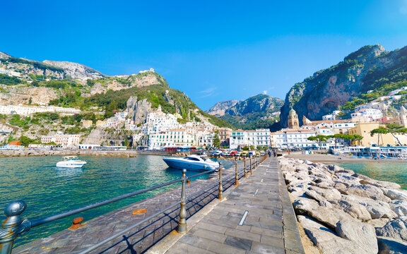Wide angle view of Amalfi harbor with boats, stone pier and coastal town below cliffs. Sunny spring day in Amalfi on Tyrrhenian coast, Campania, Italy, clear blue sky over marina, calm sea waters