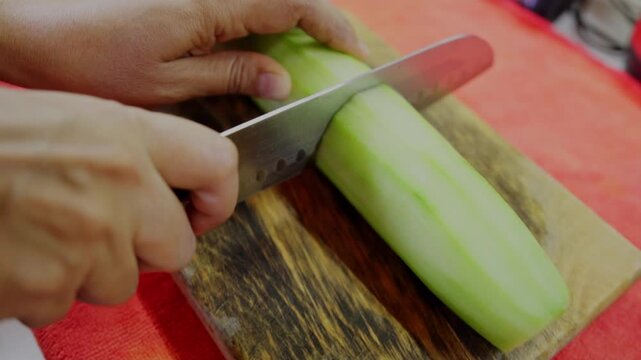 Close up of hands using a sharp knife to cut a peeled cucumber in half on a rustic wooden board, preparing ingredients for a fresh tuna ceviche.