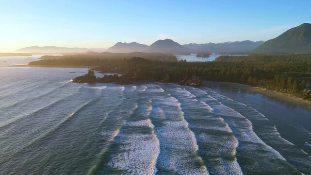 Drone shot of Cox Bay Beach, Tofino, during golden hour with waves rolling ashore, warm sunlight, and scenic mountains.