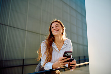 Young woman smiling and holding a mobile phone with a coffee cup, enjoying a bright urban day outdoors © we.bond.creations