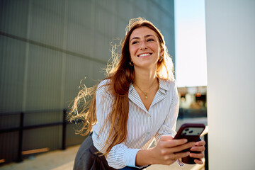 Obraz premium Young woman with freckles and long red hair smiling widely, leaning outdoors while holding a smartphone in sunlit urban setting