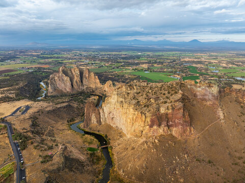 Aerial view of Smith Rock State Park with the winding Crooked River, towering basalt cliffs, and arid desert terrain under a cloudy sky in Crooked River, Oregon, United States.