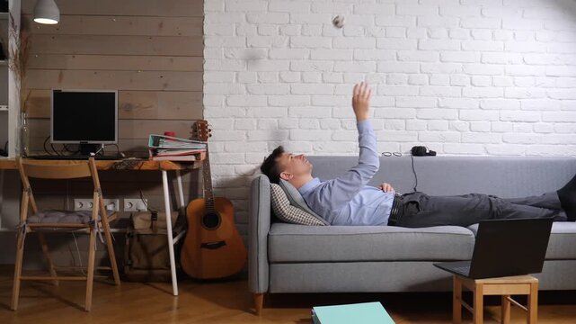 Young man procrastinating on a sofa, tossing a small ball while taking a break from his home office, laptop nearby, relaxing in casual clothes in a modern apartment living space
