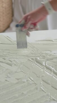 Woman spreading off-white gel, scraping with metal spatula and refining ridged texture for art