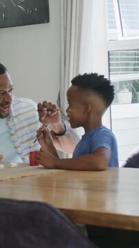 Vertical video: Leaning striped dad nudging son blue tee dipping brush, painting at table, red pot