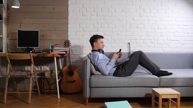 Young man multitasking on a sofa with laptop and smartphone, wearing headphones and comfortable clothes, balancing remote work, study and leisure in a cozy home office living room