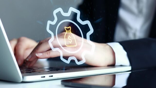 Hands typing on a laptop while looking at financial data on a screen in a modern office setting during daytime.