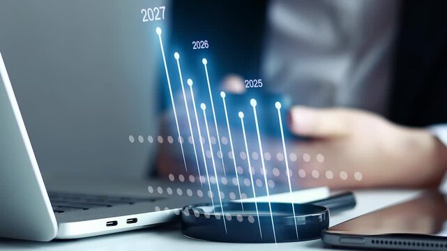 Business growth trends are shown on a screen next to a laptop in an office setting with a person handling a phone.