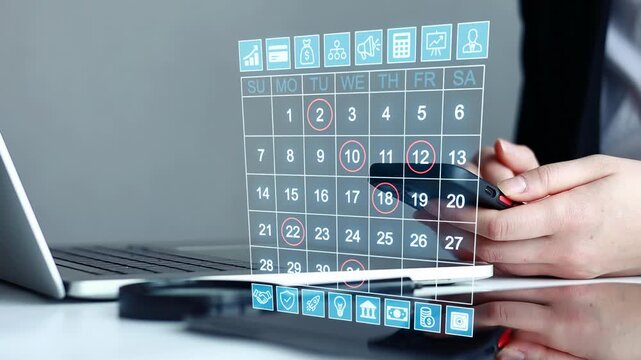 Person using smartphone with digital calendar displayed on a laptop in an office setting during daytime hours.
