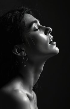 Young woman with eyes closed tilts head back in passion. Her lips are slightly parted. Soft light shines on her face and neck. She wears a delicate earring against dark background.