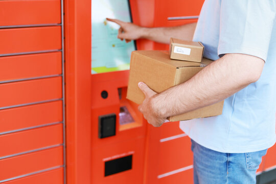 A man with cardboard boxes uses the touchscreen of an automated orange mailbox. Concept for delivery, self-service, and order pickup in e-commerce.